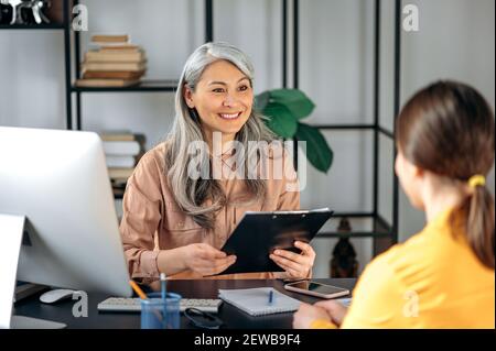 Una donna asiatica matura dai capelli grigi, datore di lavoro, esamina il curriculum di una cercatrice di lavoro femminile, seduto sul posto di lavoro. Intervista di lavoro, assunzione, ottenere un lavoro da sogno Foto Stock