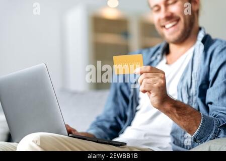 Defocused felice sorridente giovane caucasico, sta usando il laptop e la carta di credito per lo shopping in linea mentre si siede a casa sul divano. Carta di credito e laptop al centro Foto Stock