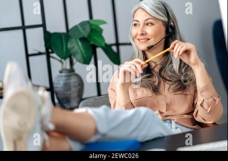 La donna asiatica anziana dai capelli grigi si siede a casa a un tavolo con le gambe gettate sul tavolo durante il lavoro, indossando cuffie, guarda via e pensando alla strategia di lavoro o alla vacanza Foto Stock