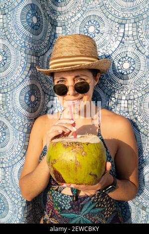 Una donna attraente in un costume da bagno, occhiali da sole e cappello che beve un cocktail di cocco fresco Foto Stock