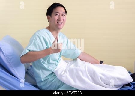 Ritratto di un giovane paziente asiatico sorridente seduto camera del letto dell'ospedale che mostra i pollici verso l'alto gesto e segno verso fotocamera Foto Stock