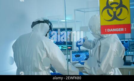 Scienziati in tuta in piedi dietro la parete di vetro che lavorano nell'area di pericolo del laboratorio. I chimici che cercano di sviluppare il vaccino usando una compressa che esamina l'evoluzione del virus ricercando per il trattamento contro il covid19 Foto Stock