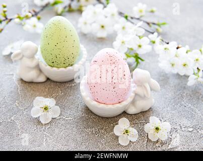 Uova di Pasqua colorate in piedi con figurine coniglietto di pasqua in ceramica e fioritura primaverile su fondo grigio in cemento Foto Stock