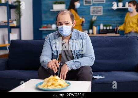 Uomo che tiene una bottiglia di birra rilassante sul divano passare il tempo con gli amici mantenendo la distanza sociale con la maschera viso in soggiorno guardando penoso alla fotocamera prevenire la diffusione del coronavirus. Foto Stock