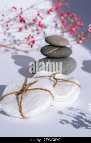 Tamponi rotondi riutilizzabili in cotone biologico per la rimozione del composto con pila di pietre di basalto su sfondo bianco. Ombre alla moda. Gypsophila o fiori rosa alito del bambino. Medicina naturale Foto Stock