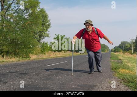 chubby ha sopportato l'escursionista anziano che si muove velocemente usando il bastone di camminare sopra Una strada nella zona rurale Ucraina in estate Foto Stock