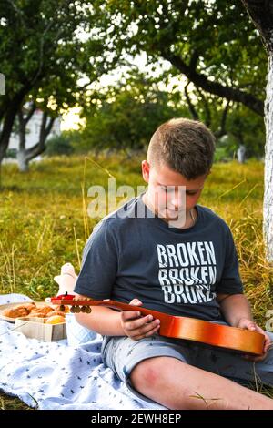 Chitarrista giovane ragazzo all'aperto. Boy on City Park estate prato godendo di una giornata a suonare la chitarra. Attività di vacanza all'aperto. Foto Stock