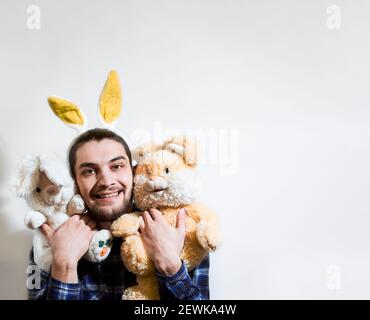 L'uomo caucasico con barba e orecchie di coniglio gialle abbraccia due conigli giocattolo imbottiti e sorride ampiamente. Banner creativo per Pasqua o negozio di giocattoli per bambini Foto Stock