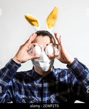 Uomo con orecchie di coniglietto pasquale. L'uomo caucasico con orecchie gialle indossa una maschera medica protettiva e tiene le uova di pollo in guscio con le mani instea Foto Stock
