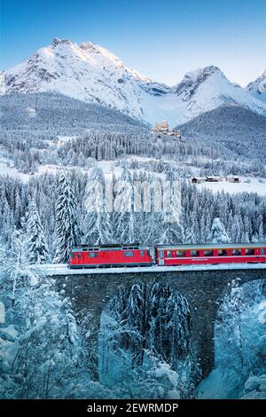 Treno Bernina Express nella foresta invernale coperta di neve che circonda il Castello di Tarasp, Engadina, Cantone di Graubunden, Svizzera, Europa Foto Stock