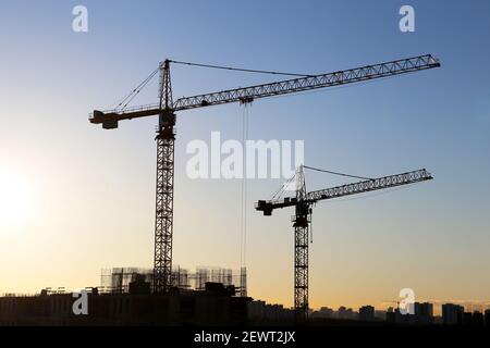 Sagome di gru da costruzione e edifici residenziali incompleti sullo sfondo dell'alba. Costruzione di alloggi, condominio in città Foto Stock