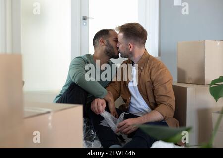 Felice multi etnico gay coppia maschile seduta circondata da scatole e baciando a casa Foto Stock