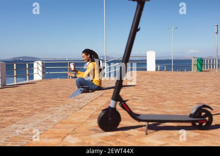 Donna afroamericana sorridente che indossa le cuffie seduto con lo smartphone passeggiata sul mare Foto Stock