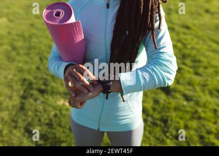 Sezione centrale della donna afroamericana che si esercita in un parco usando smartwatch e tappetino yoga per il trasporto Foto Stock