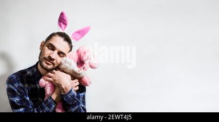 Affascinante giovane uomo caucasico con barba e orecchie rosa coniglietto pasquale abbracci ripieni coniglio stretto e chiude gli occhi in delizia. Banner con comp Foto Stock