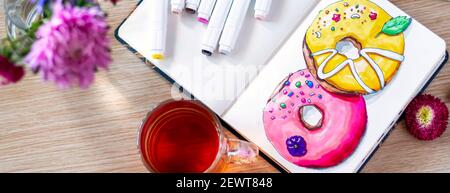Banner con ciambelle per schizzi in notebook. Il tutto posto su un tavolo di legno con una tazza trasparente di tè, taccuino, pennarelli e fiori. Hobby. Disegno don Foto Stock