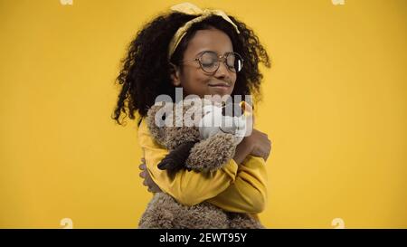 curly african american ragazza in occhiali abbracciando orsacchiotto isolato giallo acceso Foto Stock