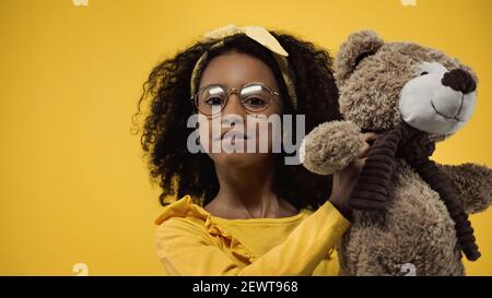 curly capretto afroamericano in vetri che tengono l'orso del teddy isolato giallo acceso Foto Stock