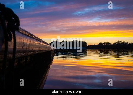 Barca galleggiante molo passerella che conduce verso il tramonto sul lago. Il sole del cielo dell'ora d'oro sotto l'orizzonte si riflette su colori vivaci e luminosi dell'acqua Foto Stock