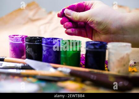 Polvere di acquerello nella mano della donna che dipinge. Vasi multicolore ad acquerello accanto agli strumenti di pittura Foto Stock