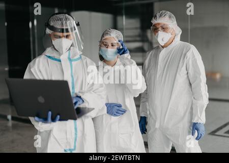 Operatori medici in tuta nocciola cercando computer portatile all'interno della clinica. Medici e infermieri con maschere protettive che parlano di picco di contagio all'interno Foto Stock