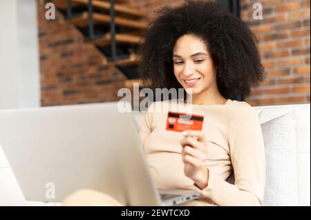 Sorridente businesswoman misto-corsa con afro hairstyle che tiene la carta di credito per fare la transazione in linea, guardando il laptop, l'acquisto degli articoli desiderati, facendo sì l'espressione della faccia, shopping dal concetto domestico Foto Stock