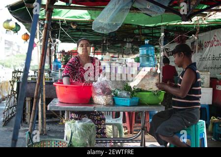 YANGON, MYANMAR - 1 GENNAIO 2020: Una donna birmana locale nel longyi tradizionale e thanaka prepara le verdure in una bancarella di cibo con un uomo birmano Foto Stock