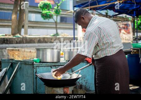 YANGON, MYANMAR - 1 GENNAIO 2020: Un uomo birmano locale nel longyi tradizionale fritta un uovo in una padella wok in un piccolo chiosco alimentare nella città centrale Foto Stock