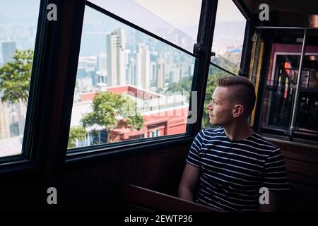 Giovane uomo che viaggia in tram fino a Victoria Peak e guardando attraverso la finestra mentre si visita Hong Kong. Foto Stock