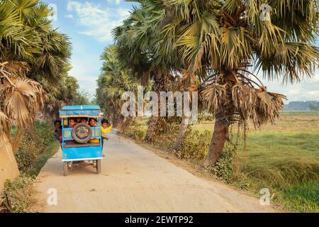 Bambini rurali in un risciò elettrico su una strada del villaggio fiancheggiata da palme in un distretto nel Bengala occidentale, India Foto Stock