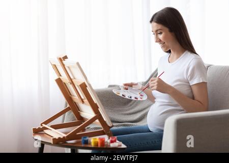 Positivo donna incinta Pittura su Easel, avendo Art Therapy sessione a casa Foto Stock