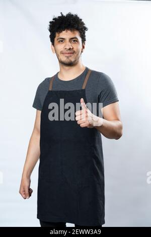 Black Apron Mockup - Ispanico Young Man in Apron Raising Thumb Up sorridente su sfondo bianco - giovane lavoratore - buon lavoro! Foto Stock