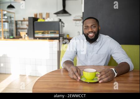 Sorridente afroamericano ragazzo è seduto nel caffè accogliente e gode di caffè, un uomo multirazziale riposa, ha una pausa, guarda la macchina fotografica allegra Foto Stock