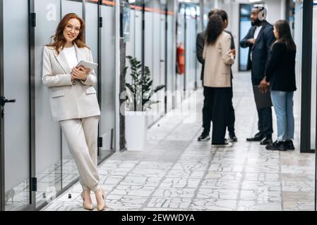 Splendida donna d'affari di successo in elegante abito elegante è in piedi nella sala d'ufficio, sorridente. Attraente dipendente rosso che guarda la fotocamera, tieni il tablet in braccio Foto Stock