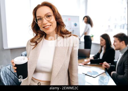 Ritratto di una giovane donna attraente rossa. Donna sicura di successo guarda direttamente il sorriso amichevole fotocamera, in background sono i suoi colleghi Foto Stock