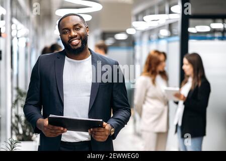 Ufficio moderno e creativo. L'uomo afro-americano sicuro, ceo, programmatore o designer, sta in ufficio guarda e sorride amichevole alla telecamera, in background i colleghi sono fuori fuoco Foto Stock