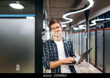 Ritratto di attraente freelance maschile in stile all'ufficio moderno. Il giovane uomo d'affari caucasico tiene il laptop in braccio, guarda la fotocamera, sorridendo, lavorando in un ufficio creativo Foto Stock