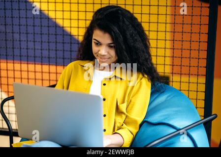 Una studentessa o un dipendente di una ragazza concentrata e soddisfatta utilizza un computer portatile, un lavoro o uno studio a distanza. Giovane donna d'affari afroamericana seduta in comoda sedia, navigando in internet Foto Stock