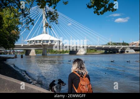 Un fuoco basso di una femmina bionda con un marrone zaino in piedi vicino a un lago con un cane sotto il luce solare Foto Stock