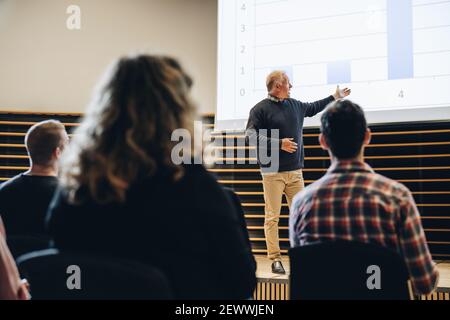 uomo d'affari che punta allo schermo di proiezione e che fa una presentazione al pubblico nel centro convegni. Relatore senior che offre una presentazione in summ Foto Stock