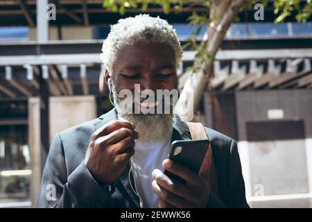 Uomo anziano afroamericano che indossa auricolari in piedi in strada utilizzando smartphone e sorridente Foto Stock