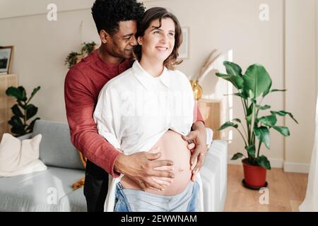 Felice uomo etnico abbracciare donna incinta a casa Foto Stock