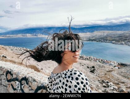 Una donna caucasica allegra in un abito voloso punteggiato di polka e. occhiali da sole con capelli soffiati dal vento Foto Stock