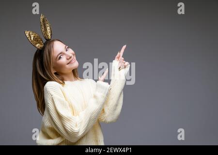 Ritratto di una bella giovane donna allegra in un maglione e nelle orecchie conigliose mostra il suo dito di lato su sfondo grigio Foto Stock