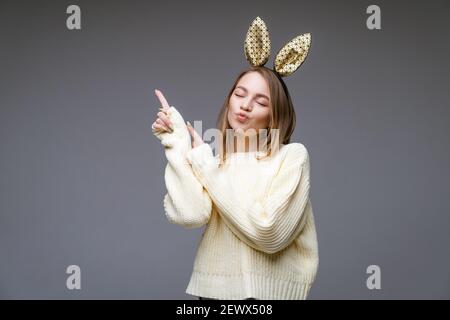 Ritratto di una bella giovane donna allegra in un maglione e nelle orecchie conigliose mostra il suo dito di lato su sfondo grigio Foto Stock