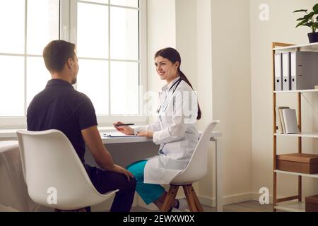 Sorridente positivo giovane donna medico seduta e prendere appunti durante visita dei pazienti dell'uomo Foto Stock