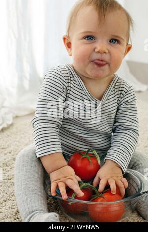 Cute baby girl sono seduti con pomodoro bianco sfondo interno. Il bambino divertente esplora le verdure Foto Stock