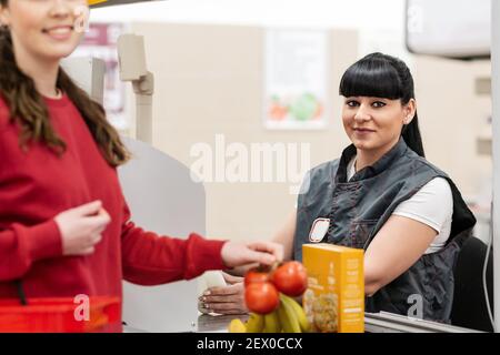 Un ritratto di giovane bella donna caucasica lavora come cassiere in un supermercato, serve clienti e posa, sorridente. Il concetto di business e tr Foto Stock