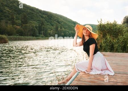 Vacanze estive. Una bella giovane donna si pone elegantemente, seduto su un molo vicino al mare e tenendo il suo cappello. Paesaggio sullo sfondo. Foto Stock