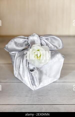 Confezione regalo trendy avvolta in tessuto di seta con tecnica Furoshiki, fiori bianchi Eustoma o Lisianthus, sfondo di legno. Concetto di durata zero sprechi. Foto Stock